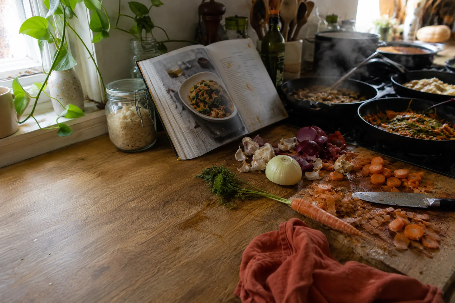 Plan de travail encombré en pleine session : livre de cuisine ouvert, épluchures, poêles occupées sur le feu